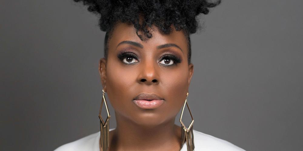 A woman looks straight at the camera with a serious faraway look. She wears a white blouse and large gold earrings. Her black curly hair is piled high on her head. 
