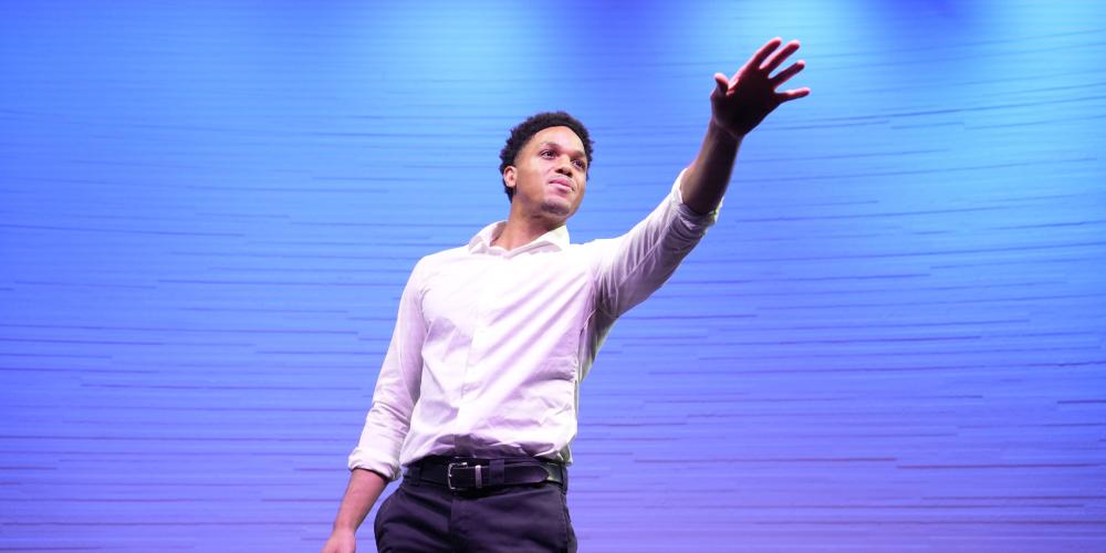 Bryce Foley on stage with light blue background and stage lights gesturing toward the audience.