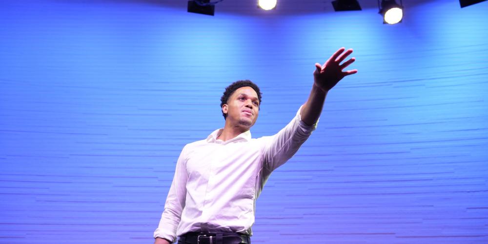 Bryce Foley on stage with light blue background and stage lights gesturing toward the audience.
