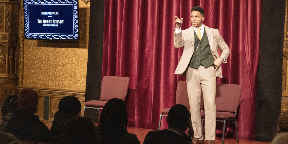 A man wearing a brown suit and green vest stands on a stage talking to the audience