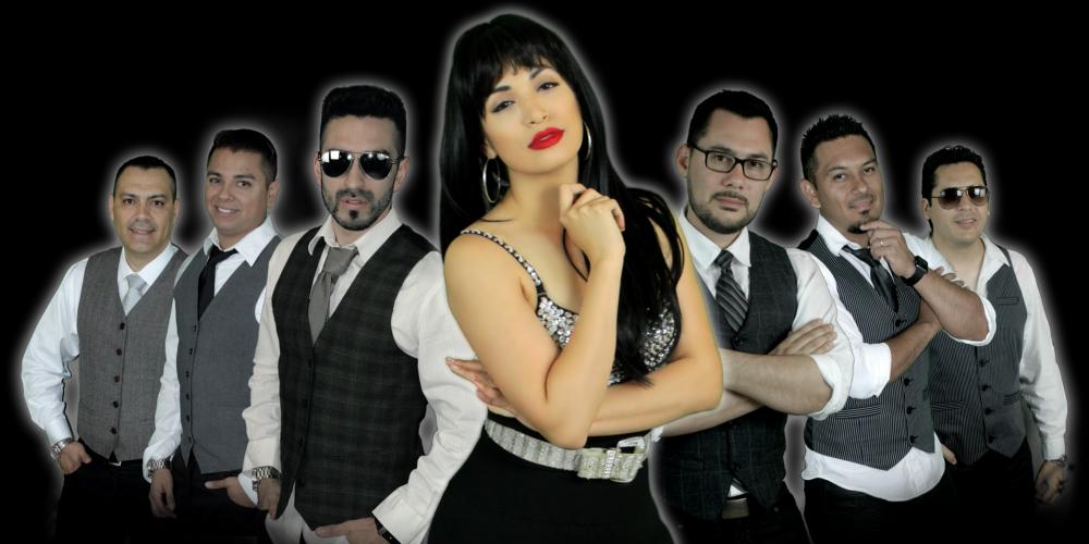 A woman with long black hair and bright red lips, wears all black with a silver studded top. Her hand is under her chin and her hip juts out to one side. Six men, three on either side of her, wear white button up shirts, black ties and vests