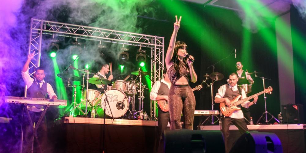 A full band plays on a stage with green and purple lights overhead. A woman stands at the front with one arm raised and a microphone in the other, singing.