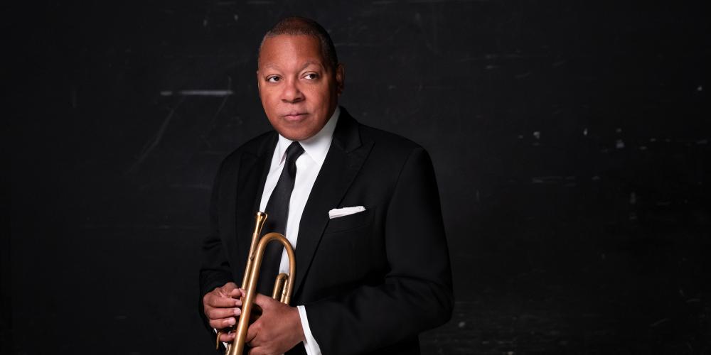 a man in a black suit stands against a black wall with a trumpet