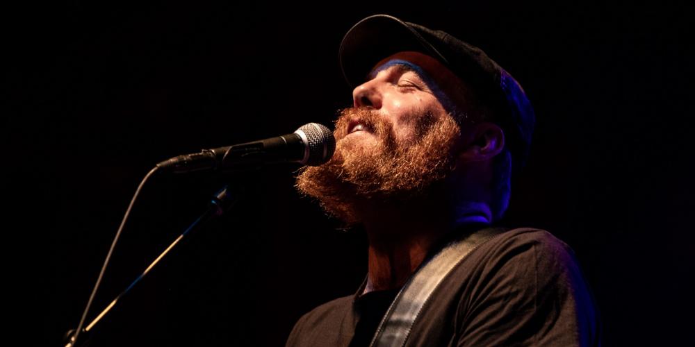 A bearded man dressed in black sings into a microphone and plays guitar