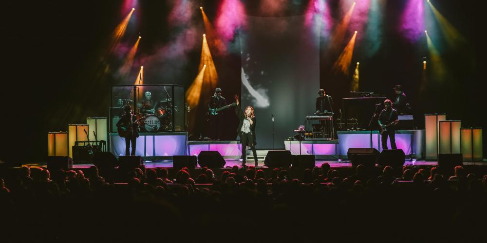A woman dressed in black raises her hand to the sky as she sings on a stage filled with colorful lights and a crowd watches