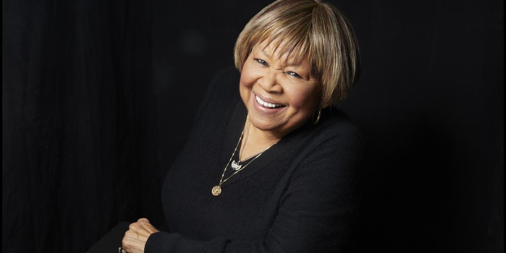 Mavis headshot with her smiling at the camera leaning on one arm and dressed in black