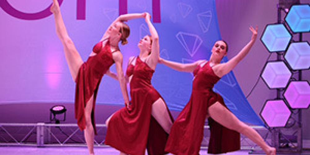 three dancers with arms and legs outstretched on stage in red dresses