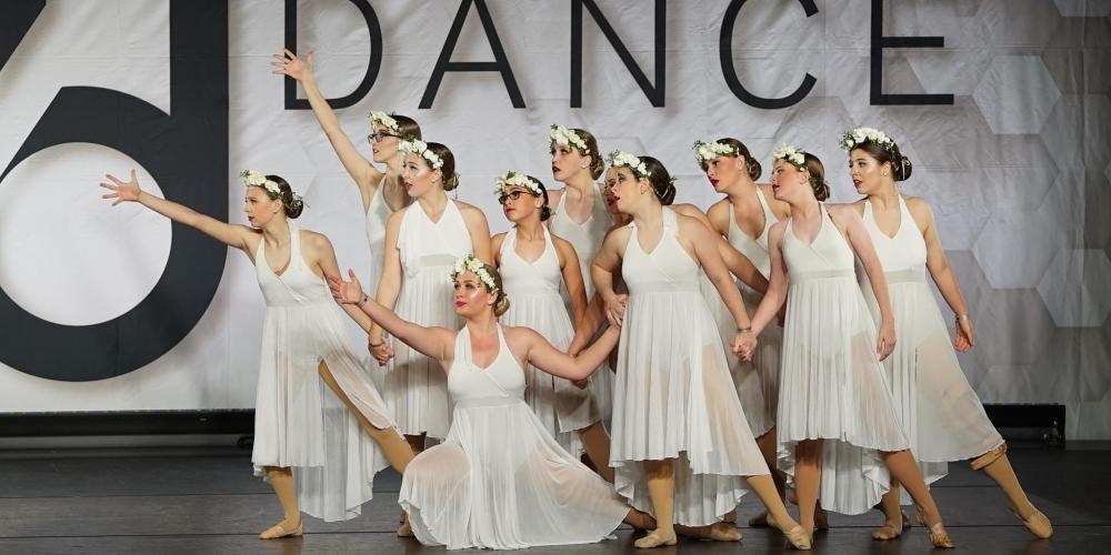 A group of barefoot modern dancers dressed in long white dresses pose on a stage