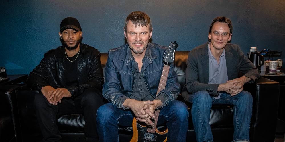 Three men sit on a couch smiling at the camera. One holds a guitar.