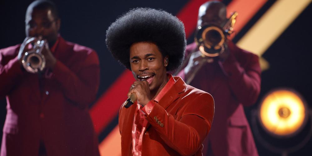 singer dressed in red singing on stage with trumpeters in background