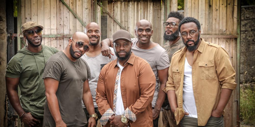 The seven male singers of Naturally 7 pose in front of a building, wearing clothes in natural color tones