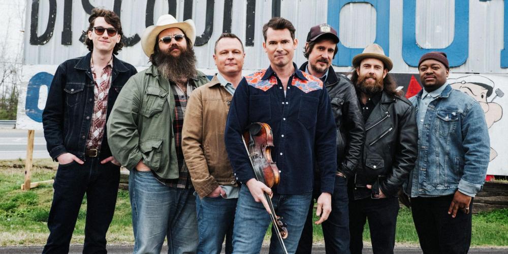 Seven men stand together in front of a sign advertising fireworks, wearing jeans and western shirts