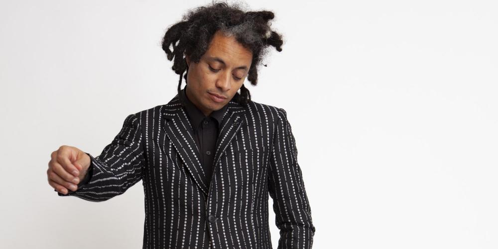 Black man with dreadlocks standing in a black suit with white pinstripes