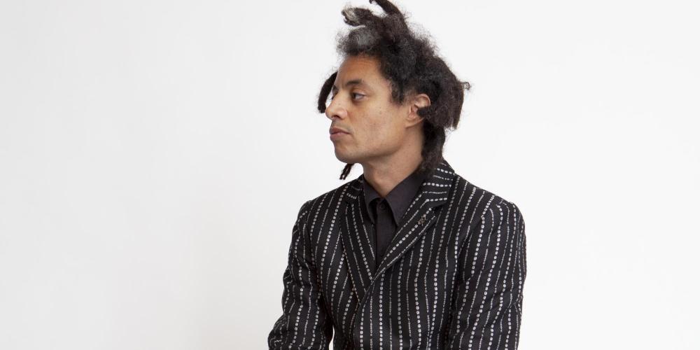 Black man with dreadlocks sitting on a box in a black suite with white pinstripes