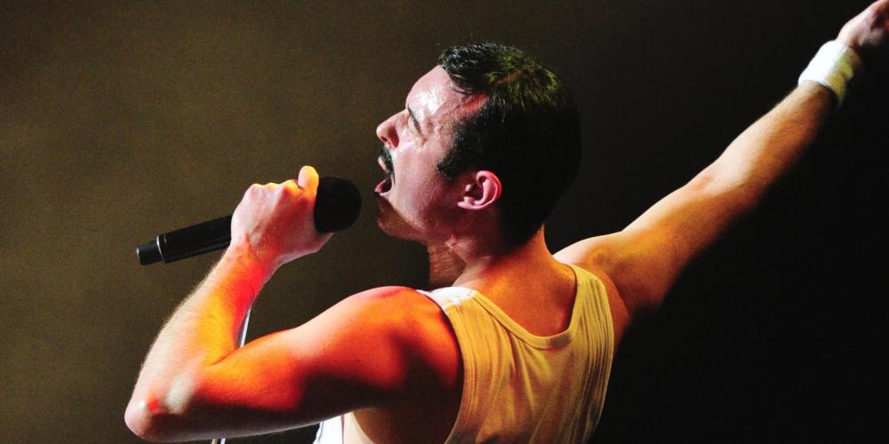 A man dressed in a white sleeveless shirt sings into a microphone with one arm raised