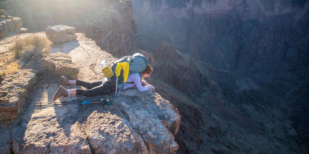 A person lays on their stomach looking over the edge of a rocky cliff, down into a canyon where the sun appears to be setting in the distance.