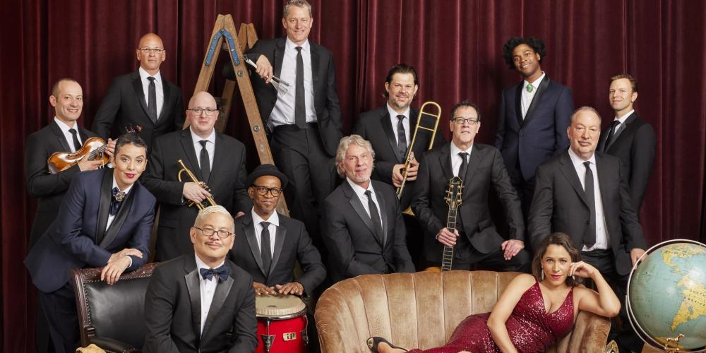 Thirteen men wearing black suites and a women in a red dress laying on her side on a sofa. 