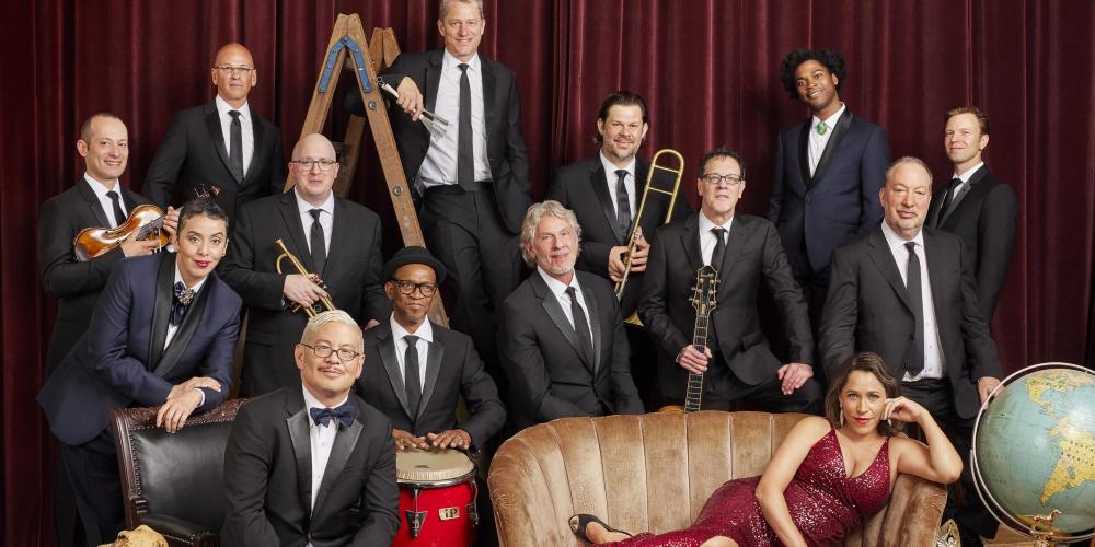14 people dressed in formal attire - 13 in tuxedos and one lounging in a red sequin dress on a couch