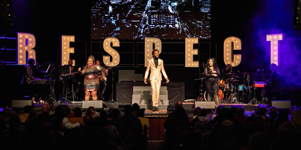 three singers performing on stage in front of large letters spelling out RESPECT