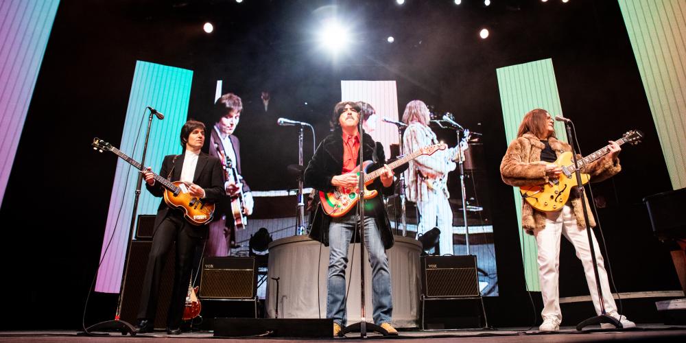 Three men wearing 60s clothing playing guitars on a stage with colorful screens behind them