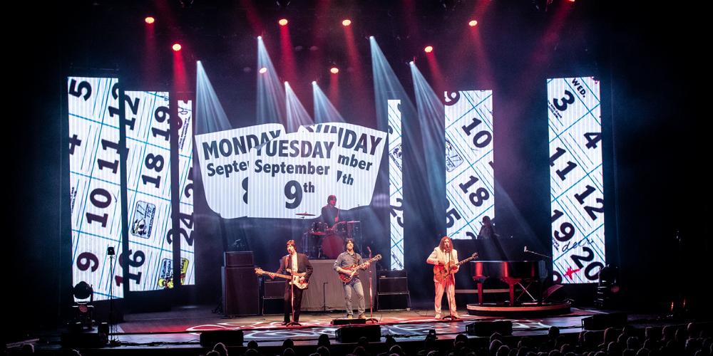 Three men dressed in clothes from the 60s stand on a stage with screens and lights behind them