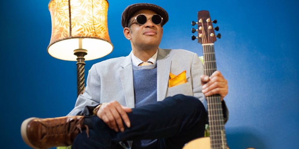a man sits in a chair in front of a bright blue wall a lamp on near his right shoulder and a guitar in his left hand. He wears dark glasses, a driving hat and a light blue blazer.