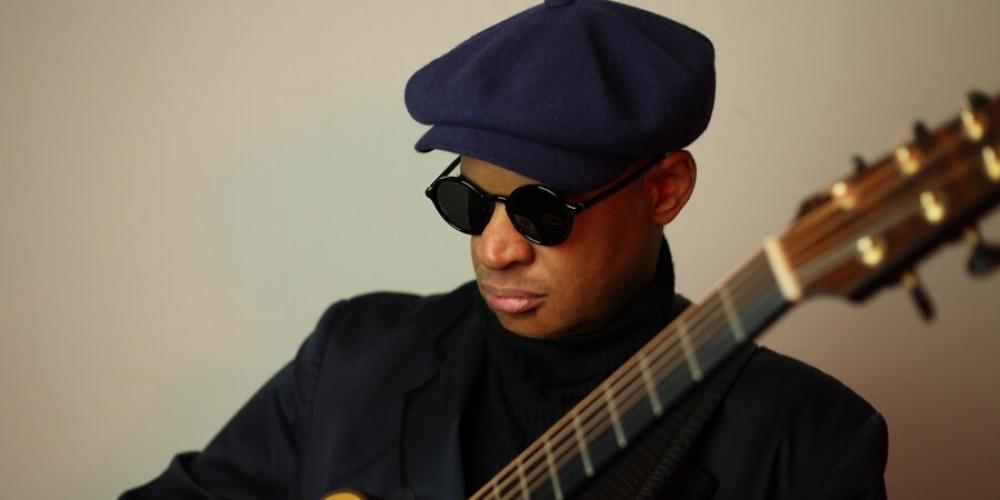 A man holds a gold colored acoustic guitar with his fingers on the fret. He wears all black with dark glasses, a turtle neck, jacket and drivers hat.