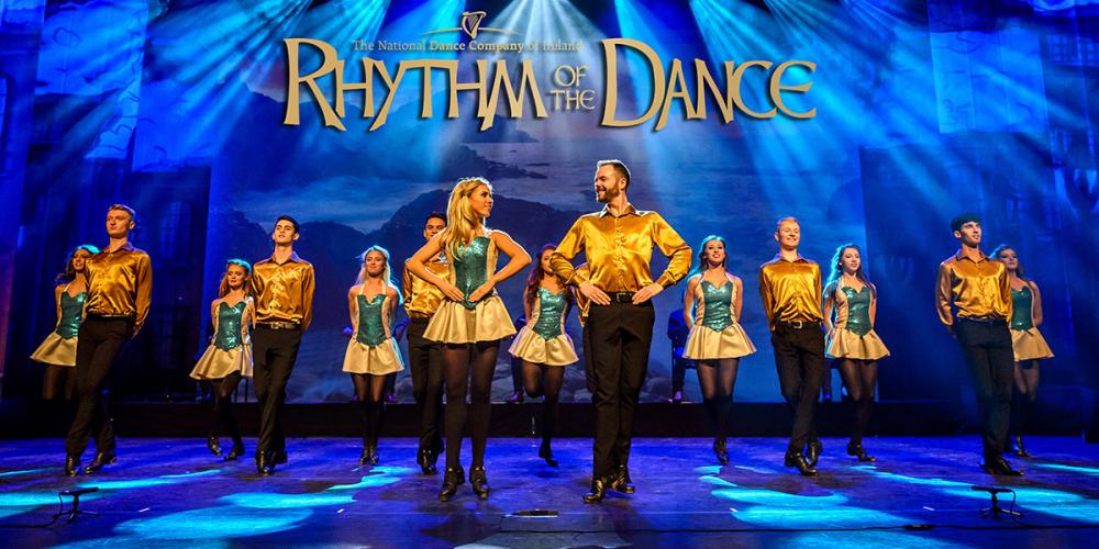 A troupe of Irish step dancers wear yellow shirts and black pants or skirts with blue lights behind them.