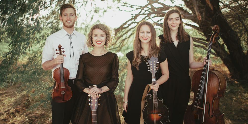 A man and three women look at the camera, string instruments in their hands with trees creating a green background