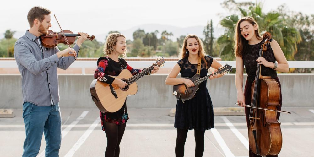 Three women and a man, sing and play their string instruments on the roof of a parking garage on a sunny day