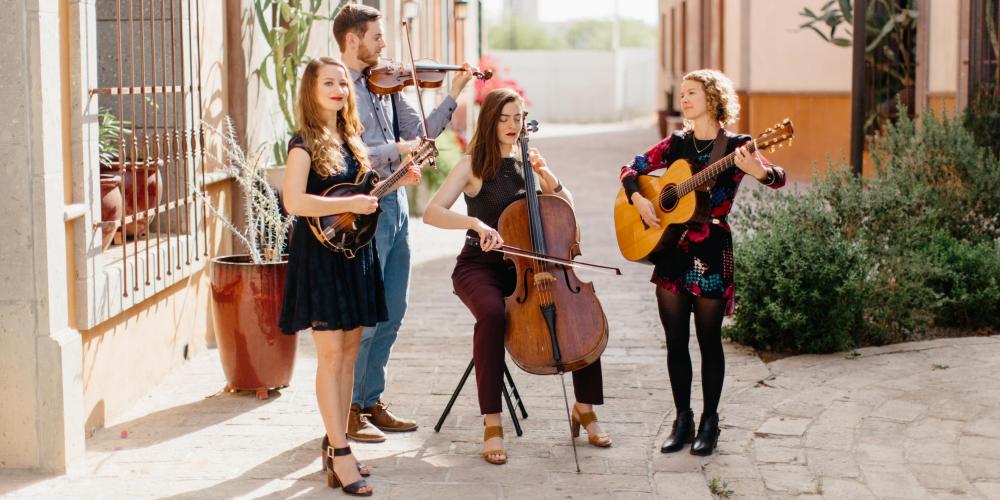 Three women and a man play their string instruments and sing in a sunny cobblestone square between adobe buildings.