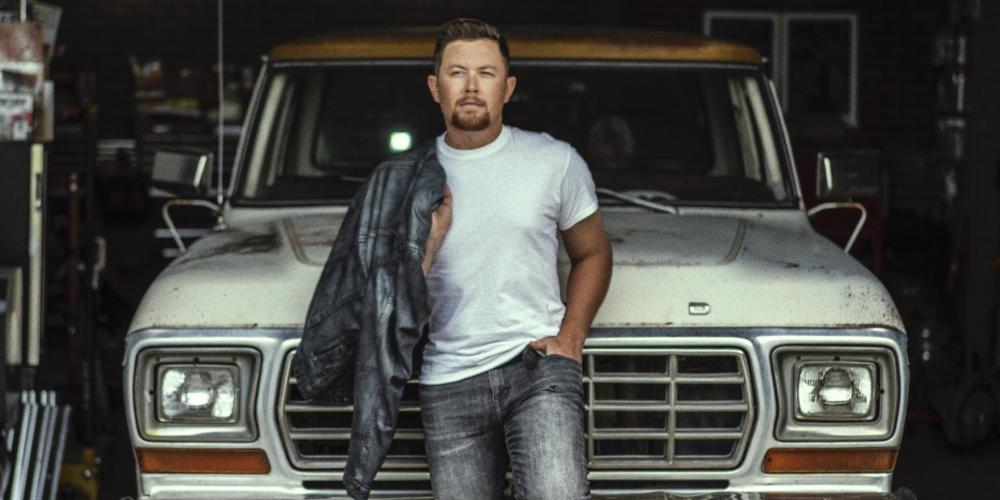 Scotty McCreery leaning back on front of an old truck wearing jeans and t-shirt