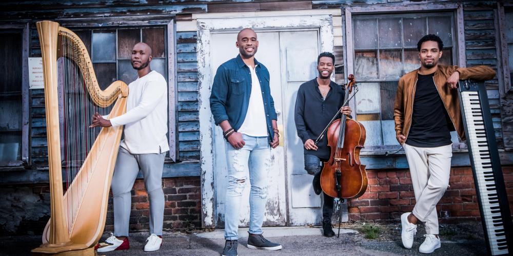 Four men stand casually in front of a weathered gray house with a white door. The main singer stands in the middle with the others gathered around with their instruments - a cello, harp and keyboard