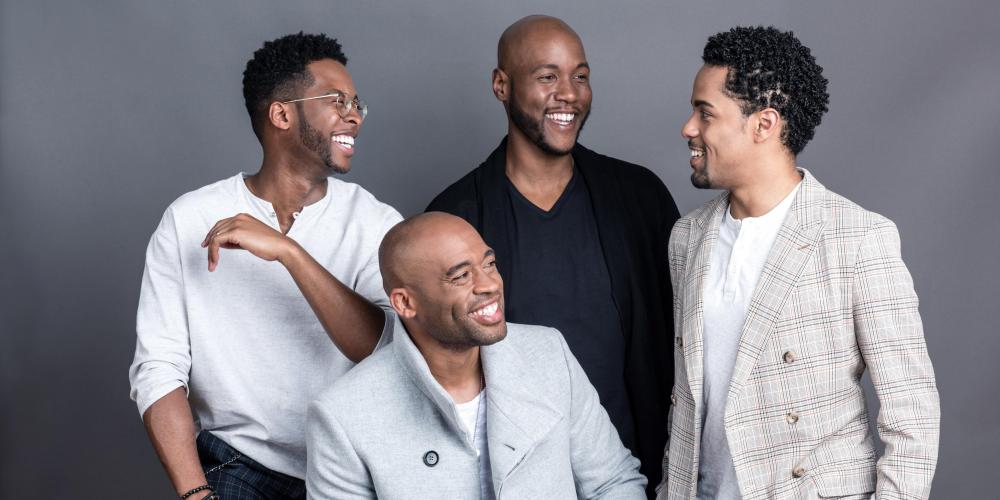 Four men pose for a formal photo with a gray background. All of them are smiling and laughing at one another, wearing gray, light blue and black. 