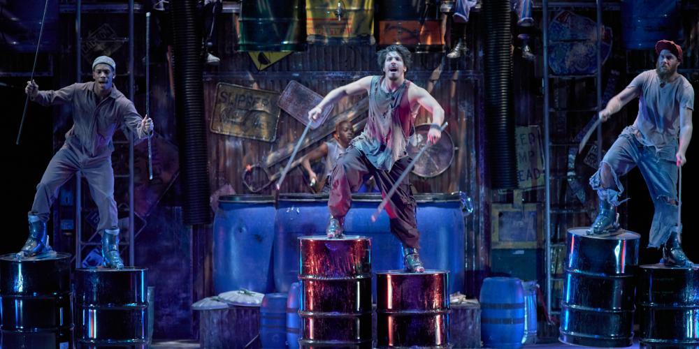 Cast members of STOMP on stage dancing with 55 gallon oil drums attached to their feet