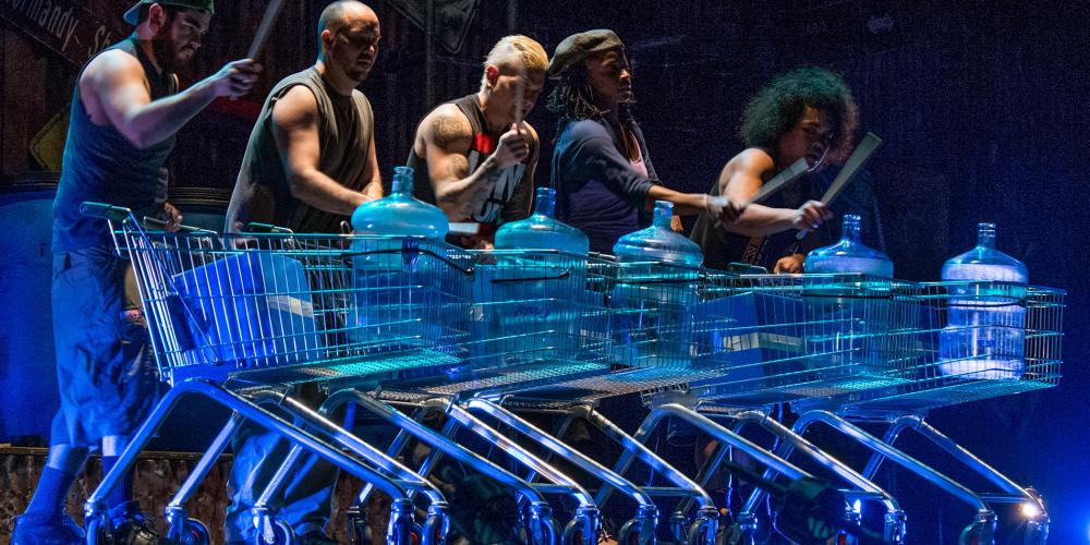 Five cast members of STOMP in a row forming a drumline playing on neon translucent drums
