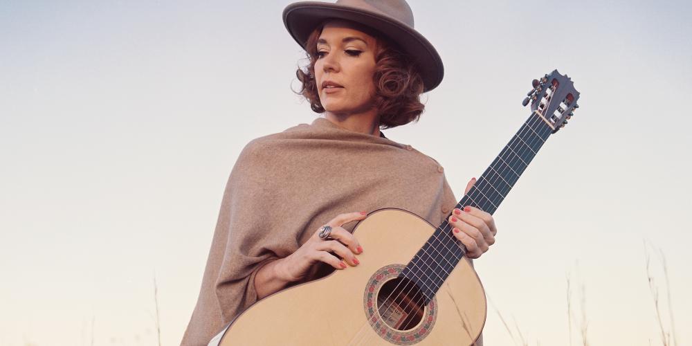 Sue Foley in field of grass looking off to the side in a hat and shall holding an acoustic guitar