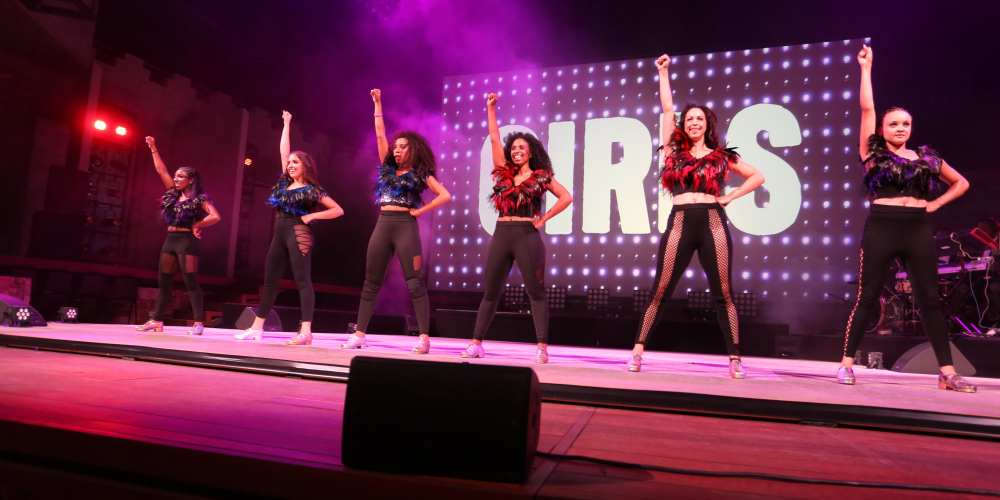Six women stand in a power pose on stage awash in purple lights with a screen behind them