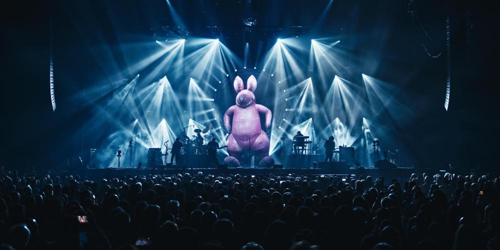A wide shot of a stage dramatically lit with blue and white lights; a giant inflatable pink bunny towers over the performers