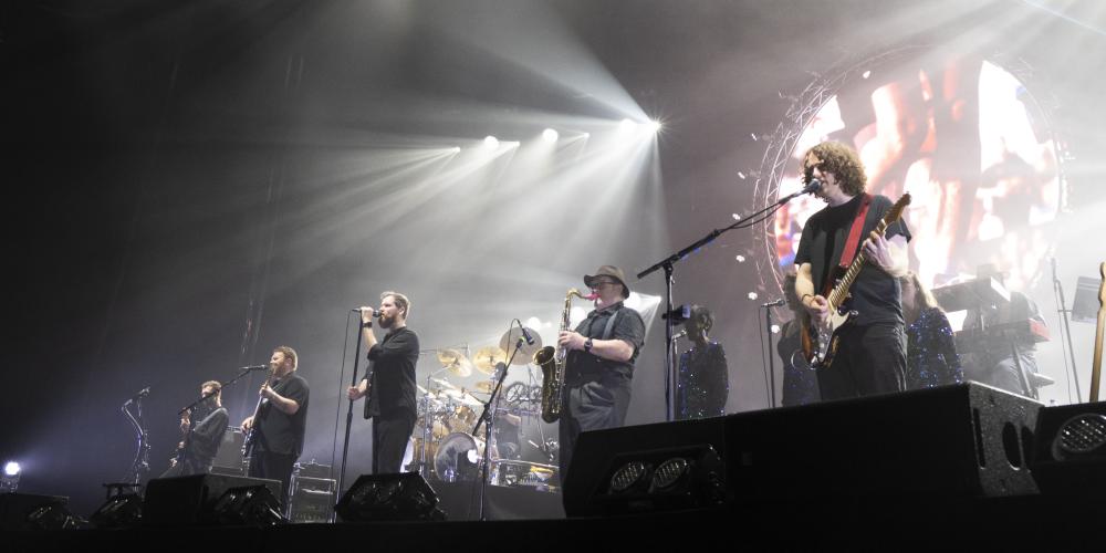 Musicians on a stage, playing instruments and singing with white spotlights and a screen behind them