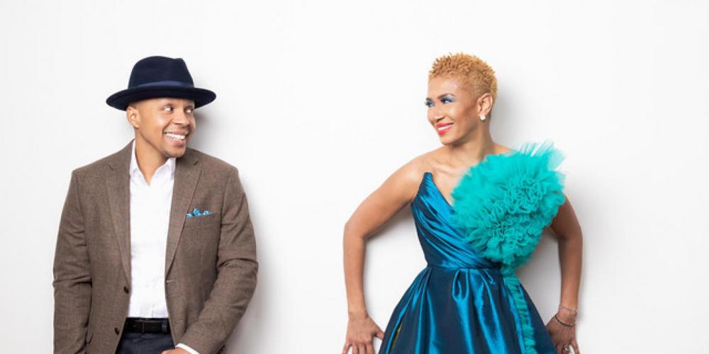 Jean Baylor and Marcus Baylor leaning against a white wall looking at each other. Jean is wearing a strapless peacock blue evening gown and John is wearing brown suit and bowler hat.