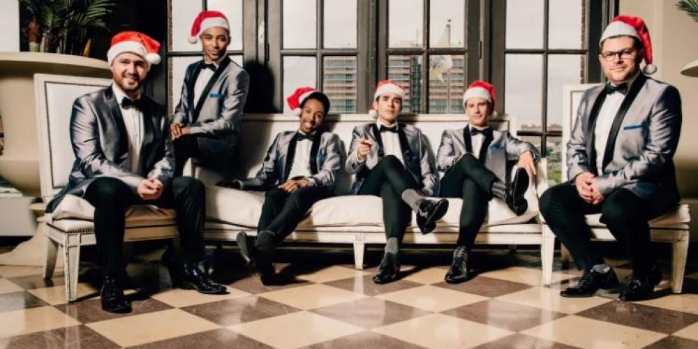 Six men wear shiny 50s style suits and red santa hats, sitting on a couch