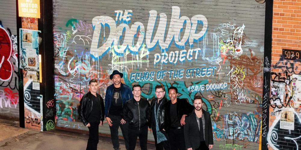 Six men standing in front  of a graffiti wall.