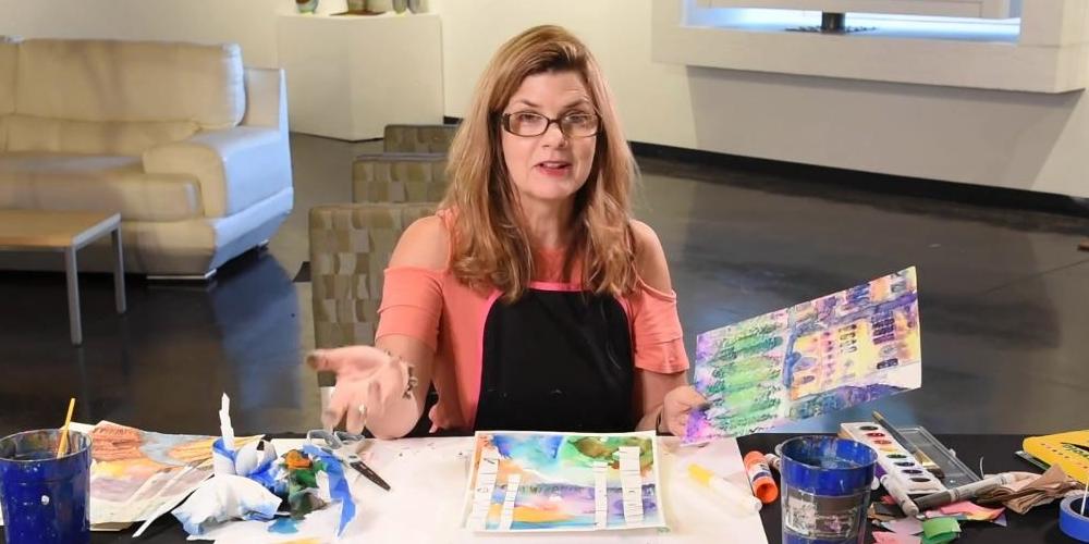 A female wearing glasses and a coral colored shirt sits at a table with art supplies and paper laid out on it