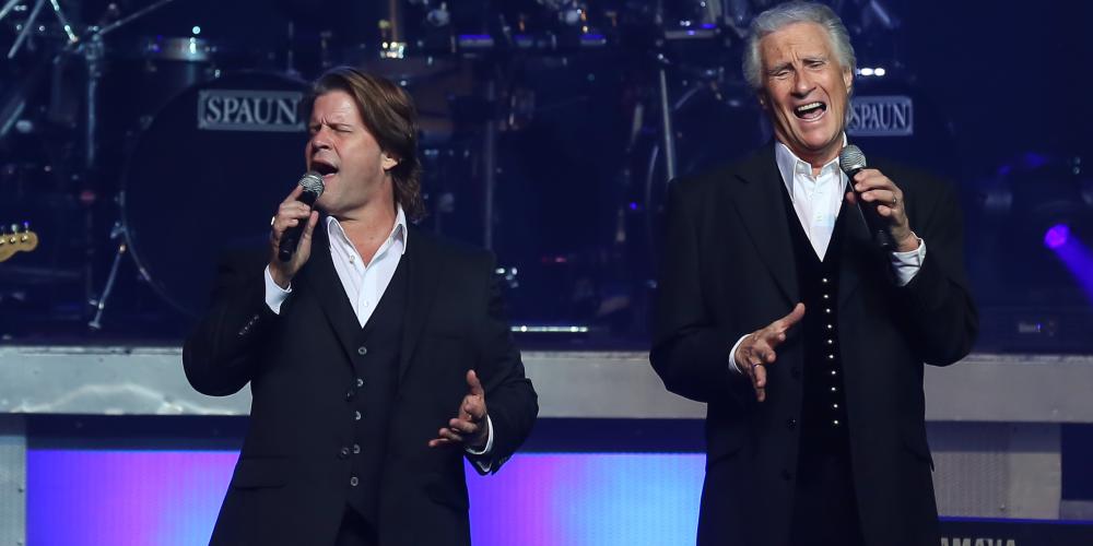 two older men in suits with gray and brown hair singing on stage