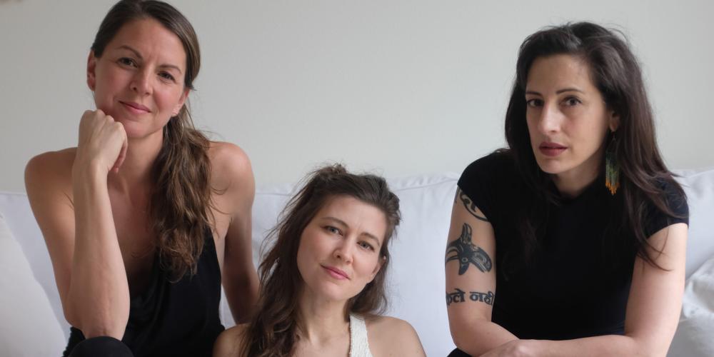 Three young women look forward at the camera with seriousness, two with brown hair and one with blond hair. 