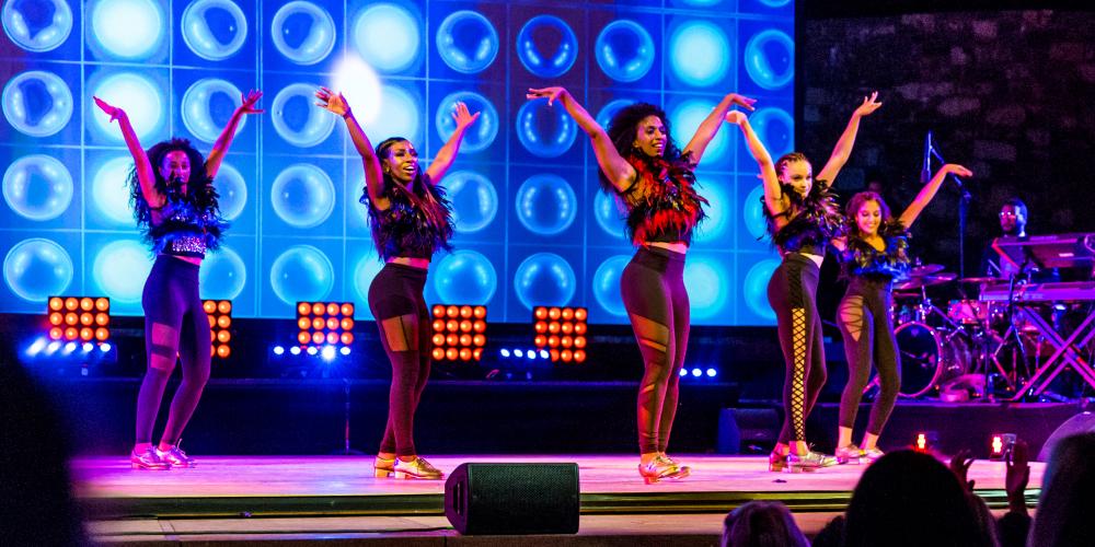 Five women dance on a stage with blue lights in the background