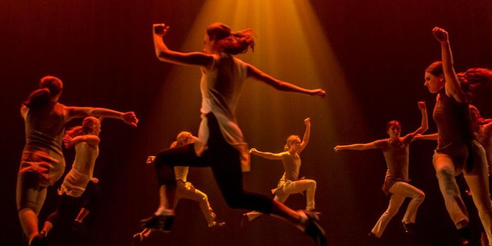 Trinity Irish Dance Company members leaping on stage in a circle with an orange spotlight shining down on them