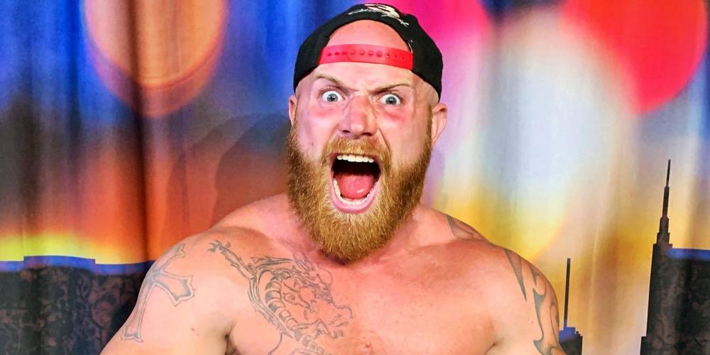 Ginger Billy wearing a baseball hat backwards, shirtless, displaying tattoos, doing a  muscle man pose holding a microphone