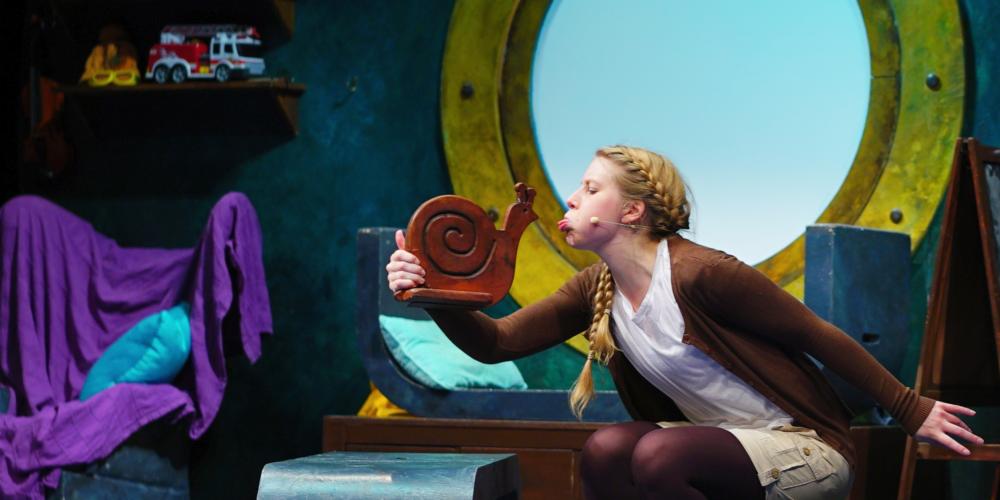 An image of a young woman sitting in a room that looks nautical, talking to a small snail sculpture in her hand